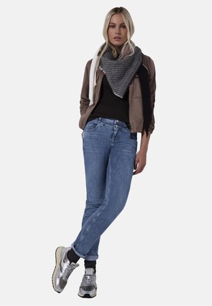 Brun bomberjakke over en sort top, blå jeans og sølvfarvede sneakers. Gråt og sort mønstret tørklæde hænger løst omkring halsen.