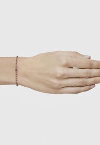 Pulsera de cuentas marrones con una gema central púrpura, llevada en una mano sobre un fondo blanco liso.