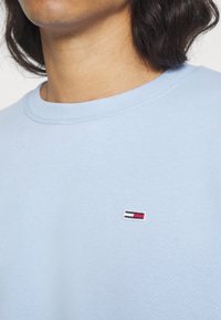 Gros plan d'une personne portant un t-shirt bleu clair à col rond avec un petit logo rectangulaire brodé en rouge, blanc et bleu marine sur la poitrine.