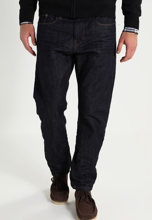 Jeans in denim blu scuro con vestibilità dritta, design classico a cinque tasche e dettagli di cucitura sottili, abbinati a scarpe in suede marrone.