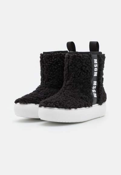 Botas de tornozelo pretas felpudas com um cabedal texturizado, sola de borracha branca e marcação preta na lateral. Possui alças para fácil calçar.
