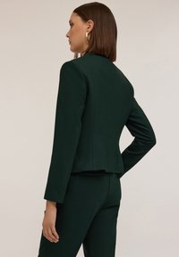 Mörkgrön skräddarsydd kavaj med strukturerade axlar, figursydd passform och en subtil textur. Har en ryggslits för ökad rörelsefrihet.