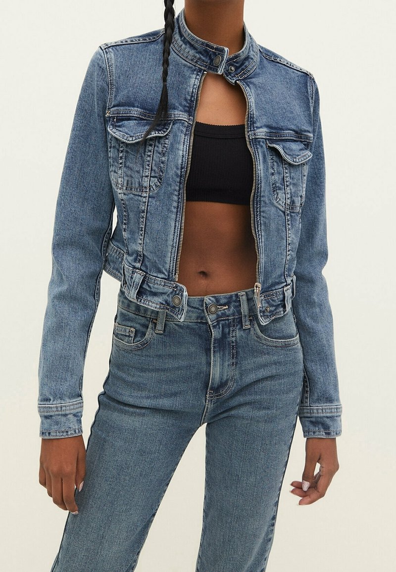 Femme portant une veste en jean bleue courte par-dessus un top noir court et un jean bleu taille haute sur un fond uni.