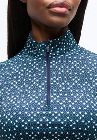 Γυναίκα που φοράει μπλε και πράσινο μπλουζάκι με φερμουάρ και ψηλό γιακά, με ορατό το άνω μέρος του στήθους και το κάτω μέρος του προσώπου.