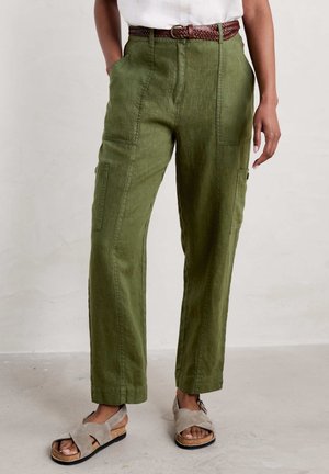 Persona que lleva pantalones de talle alto color verde oliva con grandes bolsillos, un cinturón marrón tejido, sandalias beige y una camiseta blanca parcialmente visible.