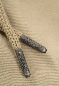 Acercamiento de extremos de cordones trenzados en beige con puntas metálicas grabadas con el nombre de la marca "Reell" sobre un fondo de tela texturizada.