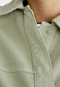 Jasnozielona dżinsowa kurtka z metalowymi zatrzaskami częściowo zapięta na czarno-białą koszulę widoczną przy kołnierzu.