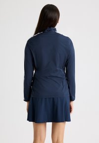 Giacca da esterno blu navy con maniche lunghe, colletto alto e vestibilità aderente. Il materiale sembra liscio e leggero.