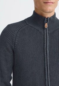 Ciemnoszara sweter z dzianiny z wysokim kołnierzem, pełnym zamkiem błyskawicznym i prążkowaną strukturą. Wyraźny skórzany akcent przy uchwycie zamka.