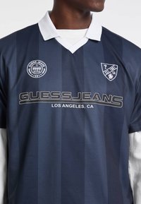 Ciemnoniebieski sweter w paski z białym kołnierzykiem. Zawiera napis "GUESS JEANS" w srebrnym kolorze oraz dwa logo: jedno okrągłe i jedno w kształcie herbu.