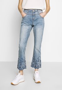 Jeans en denim bleu clair avec broderies florales à l'ourlet, bords effilochés et coupe droite ; associés à des baskets blanches.