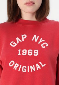 Sudadera roja con un escote redondo. Presenta el texto "GAP NYC 1969 ORIGINAL" en blanco, con bordado texturizado sobre una tela suave.