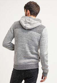 Uomo che indossa un maglione a maglia grigio con cappuccio e un motivo a strisce nere sulle spalle, mostrato da dietro, in piedi contro uno sfondo neutro.