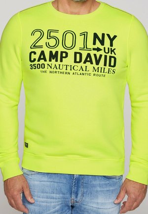 Jasno żółto-zielona koszula z długim rękawem z czarnym napisem „2501 NY → UK Camp David 3500 mil morskich Północna trasa Atlantycka”, noszona przez osobę.
