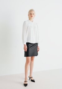 Bílá dlouhá blůza s plisováním a detaily knoflíků; v kombinaci s černou koženou mini sukni a černými jehlovými podpatky. Minimalistický design.