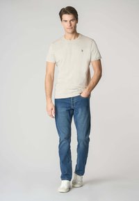 T-shirt beige a maniche corte con un piccolo logo sul petto, abbinato a jeans blu e sneakers bianche, in piedi di fronte a uno sfondo neutro.