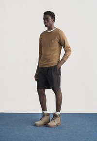 Νέος άνδρας με τα χέρια στις τσέπες, φορά καφέ μακρυμάνικο πουκάμισο, σκούρα σορτς, άσπρες κάλτσες και μπεζ ορειβατικά μποτάκια σε μπλε πάτωμα.