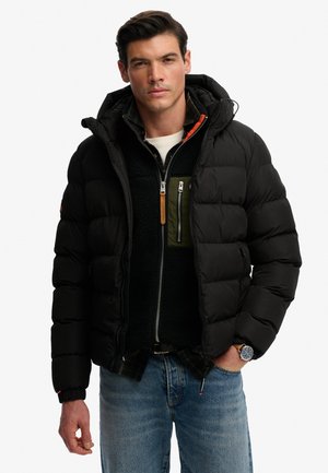 Mężczyzna w czarnej kurtce puchowej, czarnym polarku na zamek z oliwkową kieszenią, białej koszulce, niebieskich jeansach i zegarku na nadgarstku, stojący z jedną ręką w kieszeni.