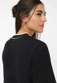Sort beklædning med en rund halsudskæring, der har kontrasterende hvid kant. Glat stofstruktur og et enkelt, tætsiddende design.