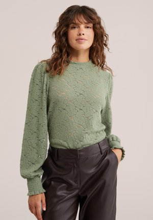 Femme aux cheveux bruns ondulés porte un haut à manches longues en dentelle vert clair et un pantalon en cuir marron foncé, une main dans la poche.