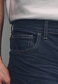 Los jeans de mezclilla azul oscuro presentan un corte clásico, costuras visibles en un amarillo contraste y un pequeño bolsillo frontal.