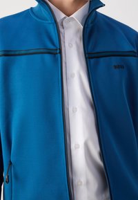 Blue zip-up jacket made of smooth fabric, featuring black accent lines, high collar, and side zippers, worn over a white button-up shirt.
