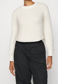 Helles cremefarbenes, geripptes Strickoberteil mit rundem Halsausschnitt und gerippten Bündchen, kombiniert mit schwarzen Cargo-Hosen. Die Textur ist weich und leicht dehnbar.