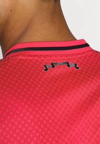 Maillot rouge avec un motif texturé, doté d'un col bordé de noir. Décoré d'un badge affichant "1975" et "2025" au dos.