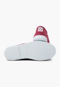 Chaussure rouge en tissu à enfiler avec une semelle en caoutchouc blanche. Elle possède un patch logo sur le talon et un motif de grip texturé sur la semelle.