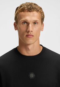 Czarna bluza z okrągłym dekoltem, heksagonalna naszywka z logo z wytłoczonymi literami na klatce piersiowej oraz gładka faktura materiału.