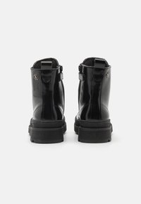 Černé lakované kožené kotníkové boty s texturovanou podešví, zadním poutkem, bočním zipem a malým kovovým logem na vnější straně.