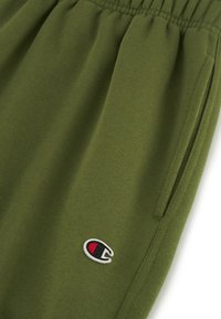 Pantalones de chándal verde oliva con cintura elástica, que cuentan con bolsillos laterales y un pequeño logo bordado en rojo, negro y blanco en el muslo.