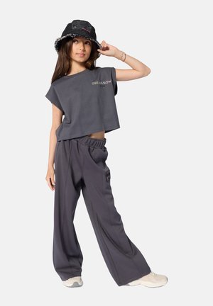 T-shirt gris court avec texte "DREAM NOW" en métal, associé à un pantalon gris ample avec une poche latérale. Chapeau bob noir avec bords effilochés.