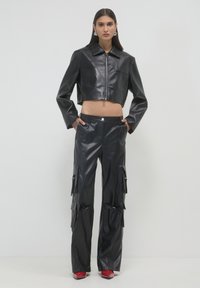 Frau in schwarzer, gefütterter Lederjacke und passenden Cargo-Hosen, stehend mit Händen in den Taschen, roten spitzen Schuhen, auf schlichtem weißen Hintergrund.