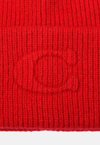 Rote gestrickte Mütze mit vertikalen Rippungen. Mit einem dezenten geprägten Logo in der Mitte. Das Material wirkt weich und texturiert.