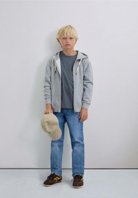 Niño rubio de pie contra una pared gris, vistiendo una sudadera con cremallera gris, jeans azules, zapatillas, sosteniendo una gorra de béisbol beige con la mano izquierda.