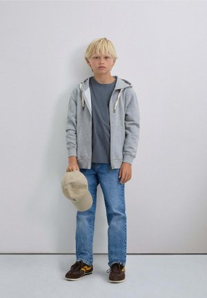 Niño rubio de pie contra una pared gris, vistiendo una sudadera con cremallera gris, jeans azules, zapatillas, sosteniendo una gorra de béisbol beige con la mano izquierda.