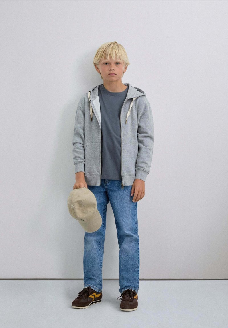 Niño rubio de pie contra una pared gris, vistiendo una sudadera con cremallera gris, jeans azules, zapatillas, sosteniendo una gorra de béisbol beige con la mano izquierda.