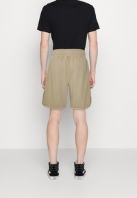 Pantalones cortos caqui con una cinturilla elástica, corte holgado y bolsillos laterales, combinados con zapatillas negras de caña alta sobre un fondo liso.