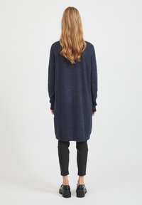 Personne aux longs cheveux blonds portant un long cardigan bleu marine, un pantalon court noir et des chaussures noires épaisses, debout devant un fond blanc.