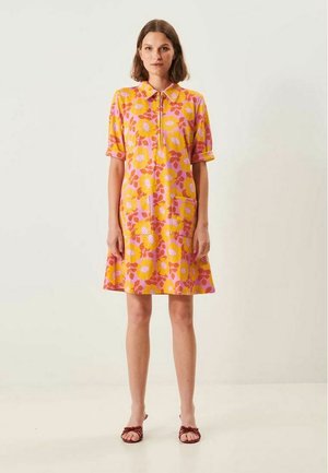 Kvinde stående iført en knælængde gul kjole med lyserøde blomsterprint, korte ærmer, krave, forlommer og brune sandaler.