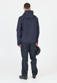 Marinblå vattentät jacka med huva och kontrasterande svarta sidopanels, i kombination med svarta vattentäta byxor. Personen håller en svart hjälm.