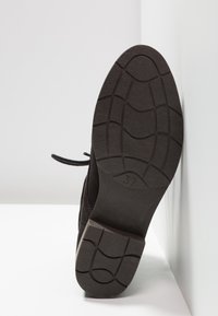 Semelle d'une chaussure noire à lacets avec une semelle à motifs et le numéro de taille 37 visible, positionnée verticalement contre un coin blanc.