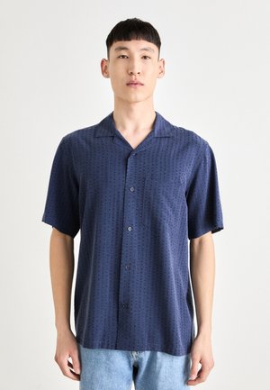 Kortermet skjorte med knapp i mørk blått stoff med subtilt mønster, lomme på venstre bryst og rett fald, kombinert med lyse denim.