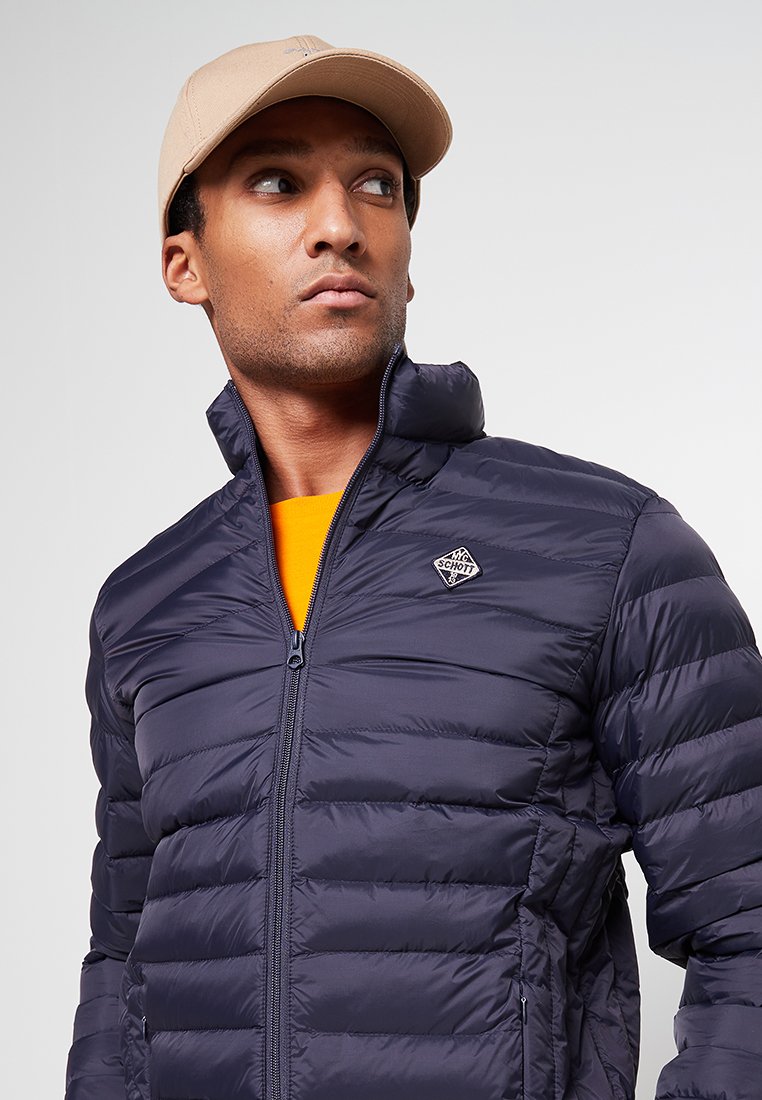 Marineblauer Winterjacke mit hohem Kragen, Reißverschluss und horizontalem Steppmuster. Mit Logo-Patch auf der linken Brust. Oranges Innenfutter.