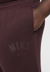 Maroon atletiska joggers med elastisk midja; har en subtil logotyptryck i mörkare maroon på sidan och en sidoficka.