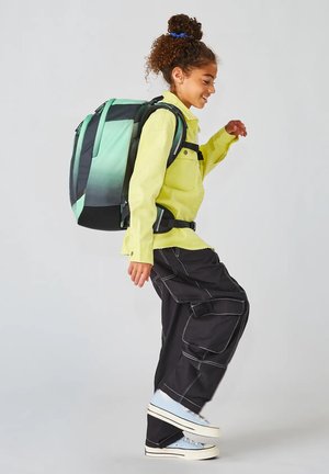 Uno zaino con una sfumatura di nero e verde indossato da un bambino, abbinato a una maglietta gialla brillante e pantaloni cargo neri con cuciture bianche a contrasto.
