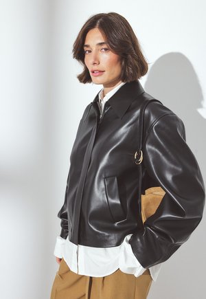 Donna che indossa una giacca di pelle nera sopra una camicia bianca, pantaloni beige e porta una borsa in suede marrone a tracolla.