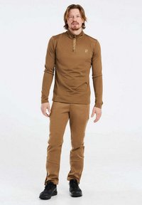 Brun långärmad topp med halvdragkedja och logotyp, tillsammans med matchande bruna byxor, gjorda av ett mjukt, töjbart tyg. Svarta stövlar.