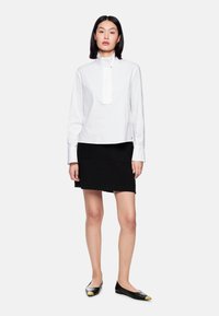 White long-sleeve blouse with a stand collar and front tie, paired with a black textured mini skirt featuring two front pockets and flat shoes.